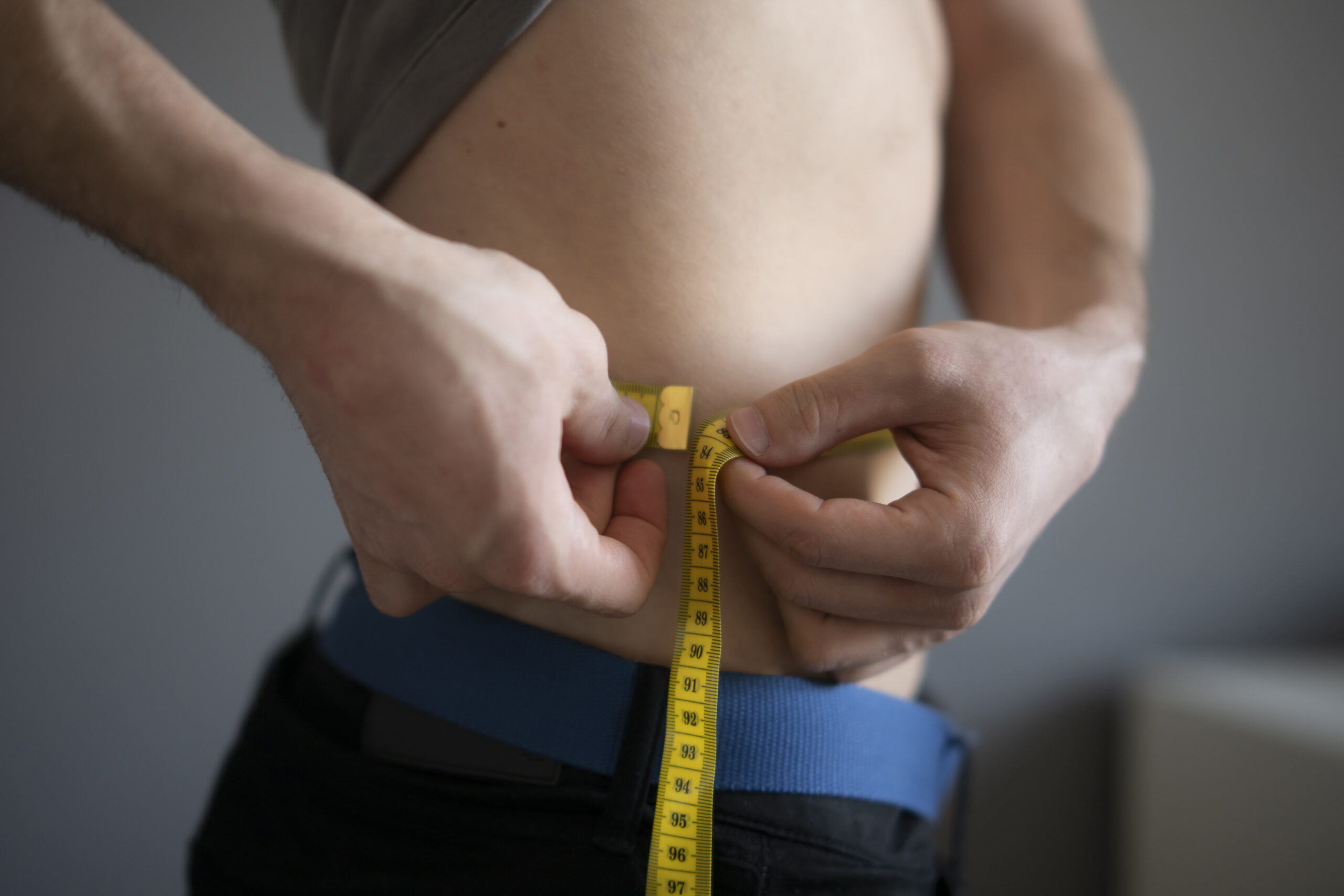 A close up view of a man holding a yellow measuring tape around his waist, checking his measurements possibly as part of a fitness routine or weight loss program The focus is on his hands securely holding the tape at the naval level, highlighting the numb